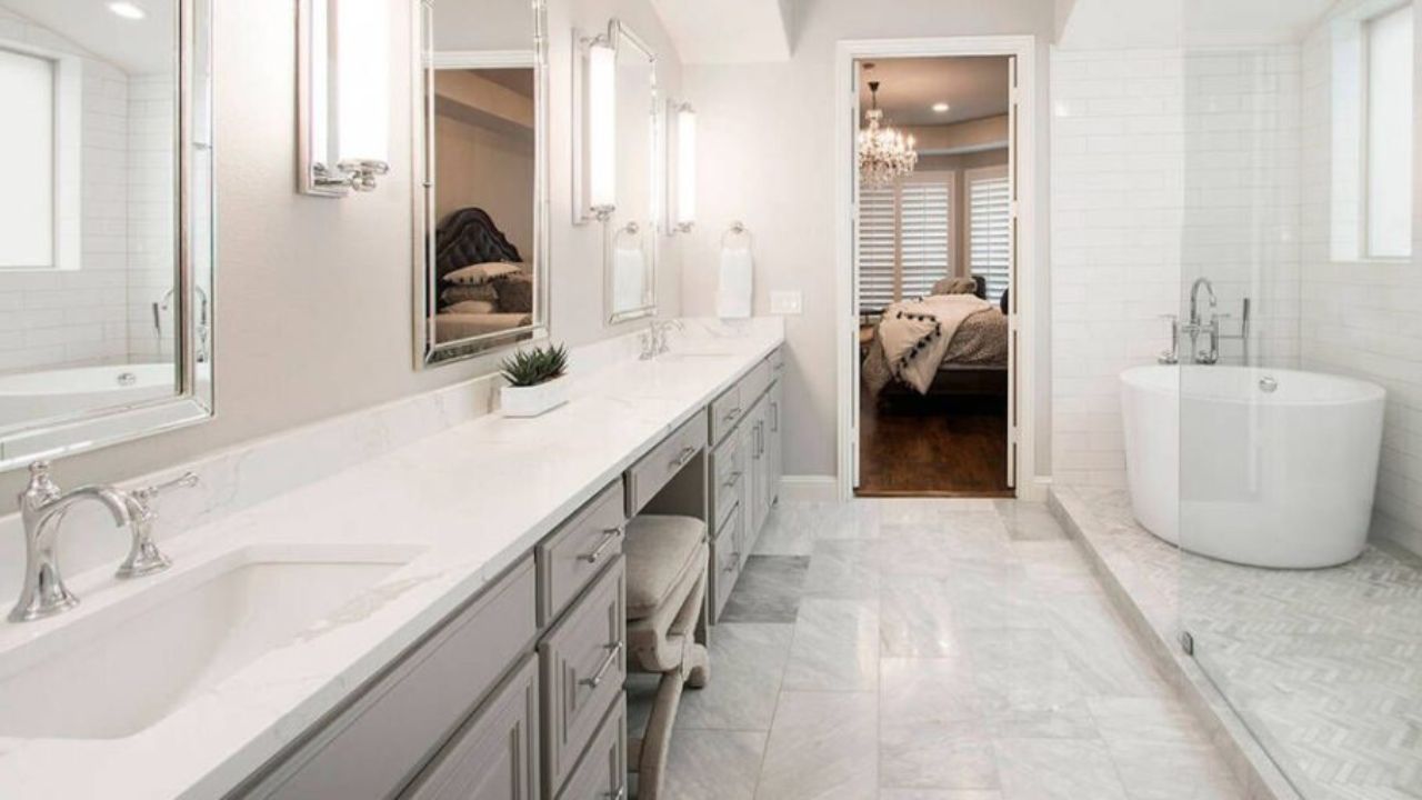 Spacious and modern bathroom featuring sleek gray cabinetry, marble flooring, a freestanding bathtub, and large mirrors. The design reflects expert bathroom remodeling with high-quality finishes and a seamless transition to a well-lit bedroom.