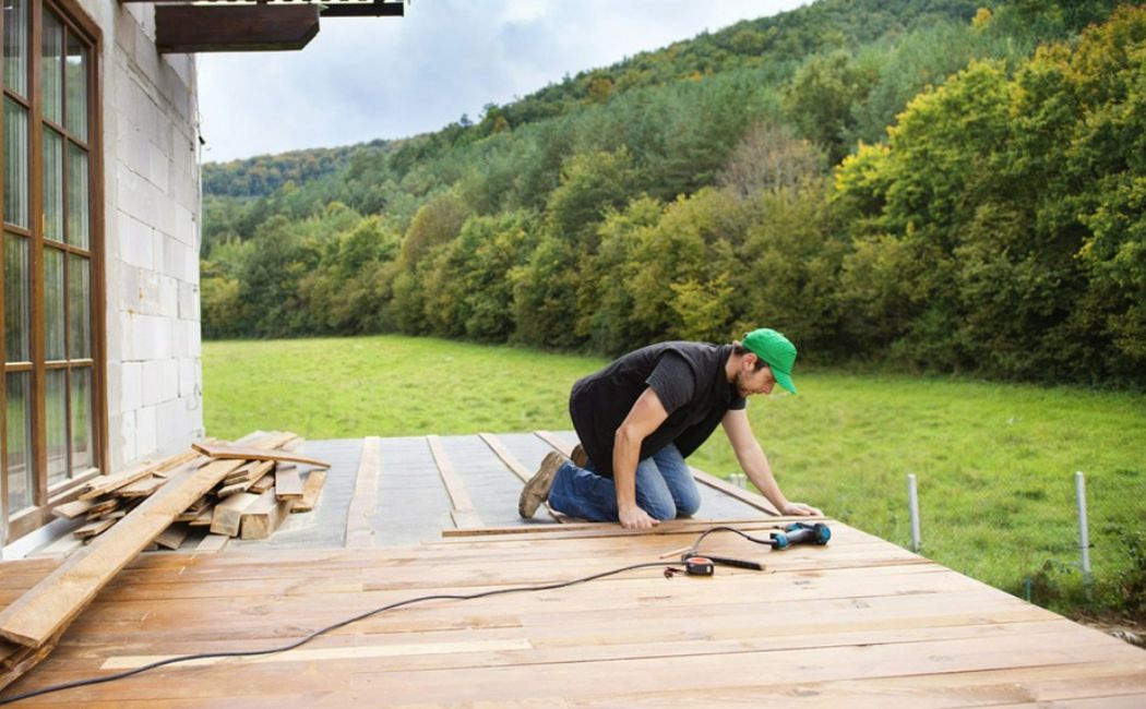 A professional carpenter installing a wooden deck outside a house surrounded by green hills and trees, showcasing expert Deck Installation Services in a scenic rural setting.