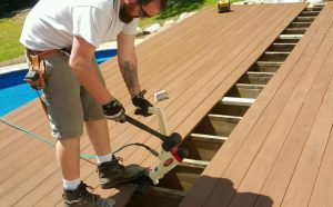 A professional contractor installs composite decking boards using a specialized tool during a poolside deck construction project. The scene highlights skilled craftsmanship and precision, showcasing high-quality deck building services in progress.