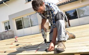 A skilled worker uses a power drill to secure wooden planks during the construction of a backyard deck in front of a modern home. The image showcases professional deck installation services, emphasizing attention to detail and quality craftsmanship.