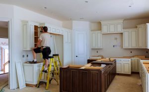 A skilled worker is seen installing kitchen cabinets in a newly renovated space. The kitchen features elegant white cabinetry with a dark wooden island, reflecting high-quality craftsmanship. The area is still under construction, with some cabinets missing doors and tools scattered across the floor. Representing best remodeling services.