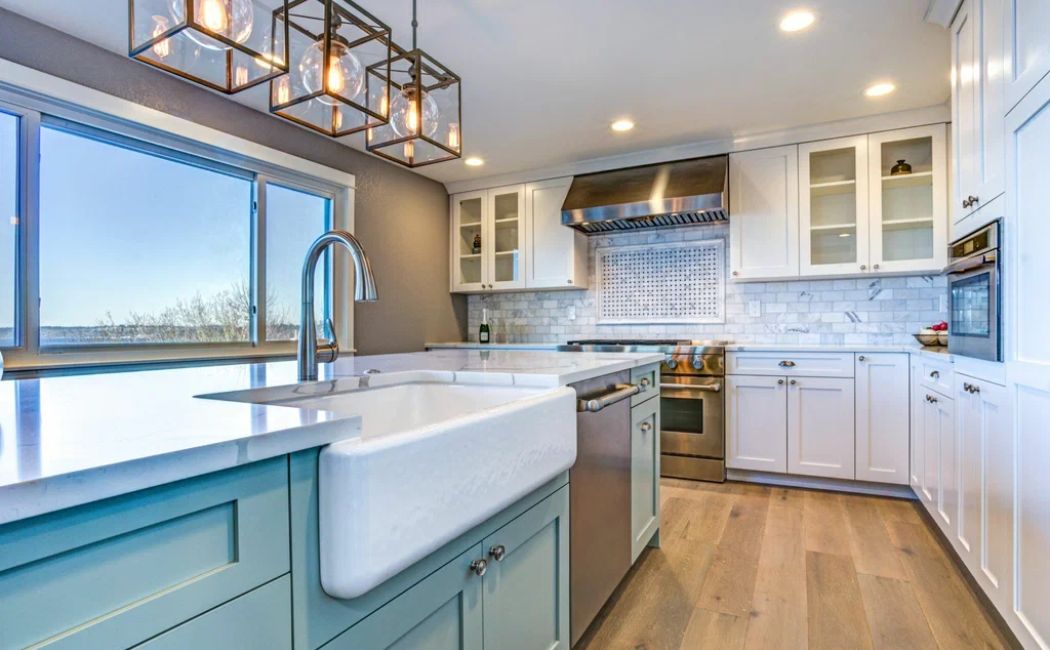 A modern kitchen with white cabinetry, stainless steel appliances, and a farmhouse sink showcases the elegance and functionality achieved through professional Remodeling Services. The space features a marble backsplash, wooden flooring, and contemporary lighting, blending style and practicality for an inviting cooking environment.