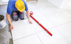 A construction worker wearing a yellow helmet installs large ceramic tiles on a floor, using adhesive and a level tool for precision, part of professional flooring services for residential or commercial spaces.