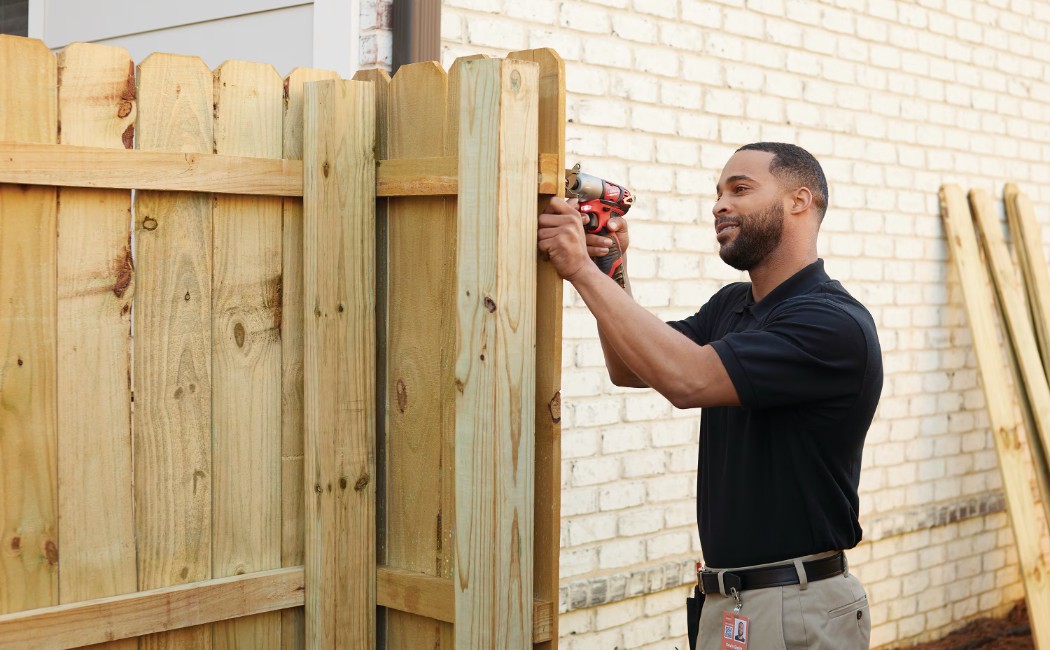 Professional Fence Repairing Services for Residential Properties A professional technician repairing a wooden fence using a power drill beside a brick house, showcasing reliable Fence Repairing Services for residential properties.