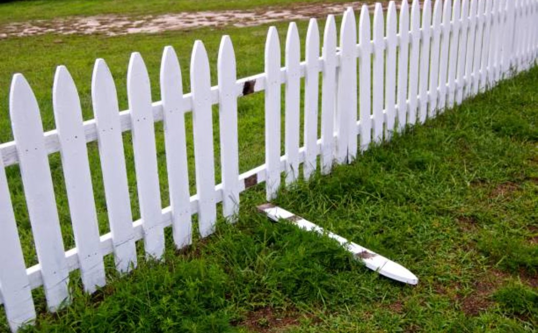 Damaged white picket fence with a broken panel lying on the grass, highlighting the need for professional Fence Maintenance Services to restore safety and appearance.